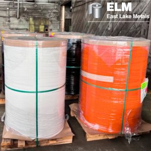 Large, shrink-wrapped vertical coils of white and orange architectural steel on wooden pallets, demonstrating the raw material quality required for superior prepainted metal color retention at East Lake Metals.