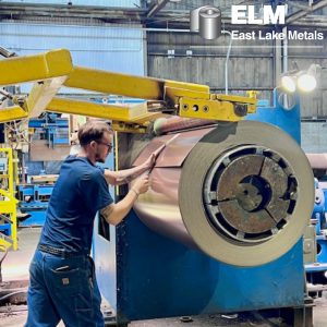 East Lake Metals, a trusted metal supplier, showing an experienced technician meticulously inspecting a large copper-colored architectural metal coil as it is processed on an industrial uncoiler machine for precision fabrication.